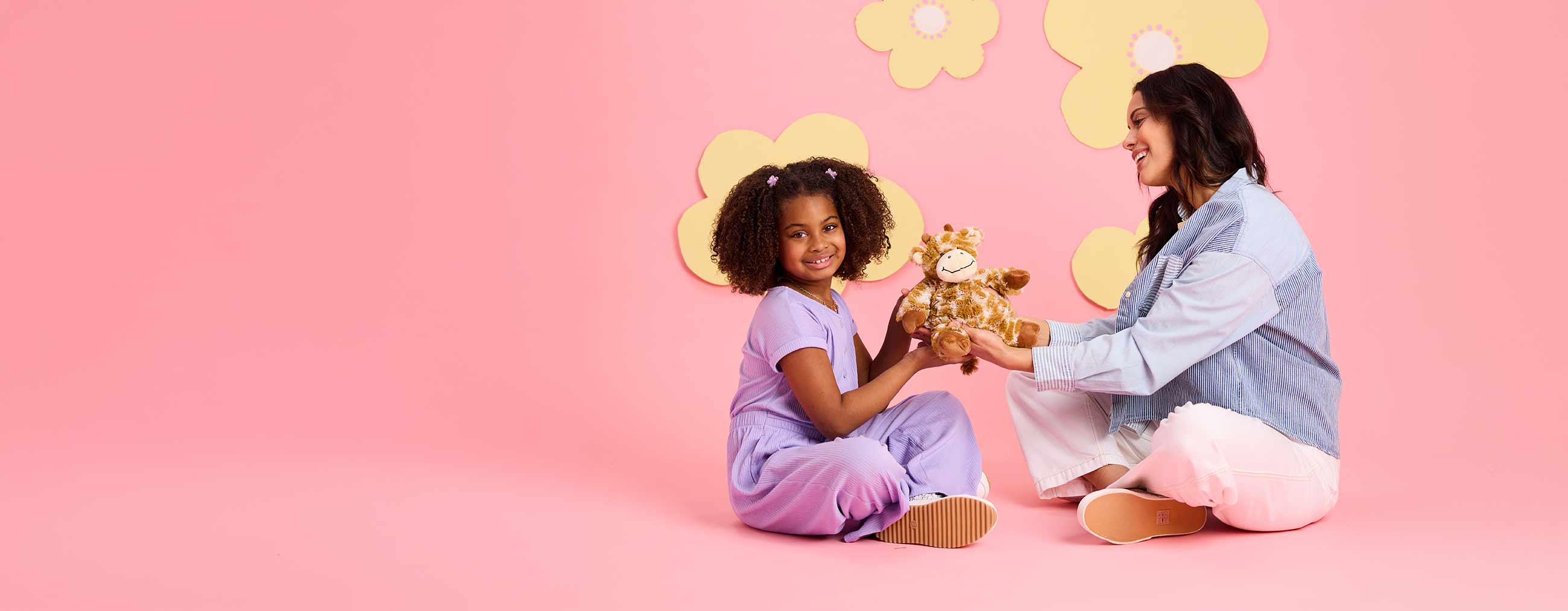 Woman and child sitting on a pink floor with a teddy bear, surrounded by yellow flowers.
