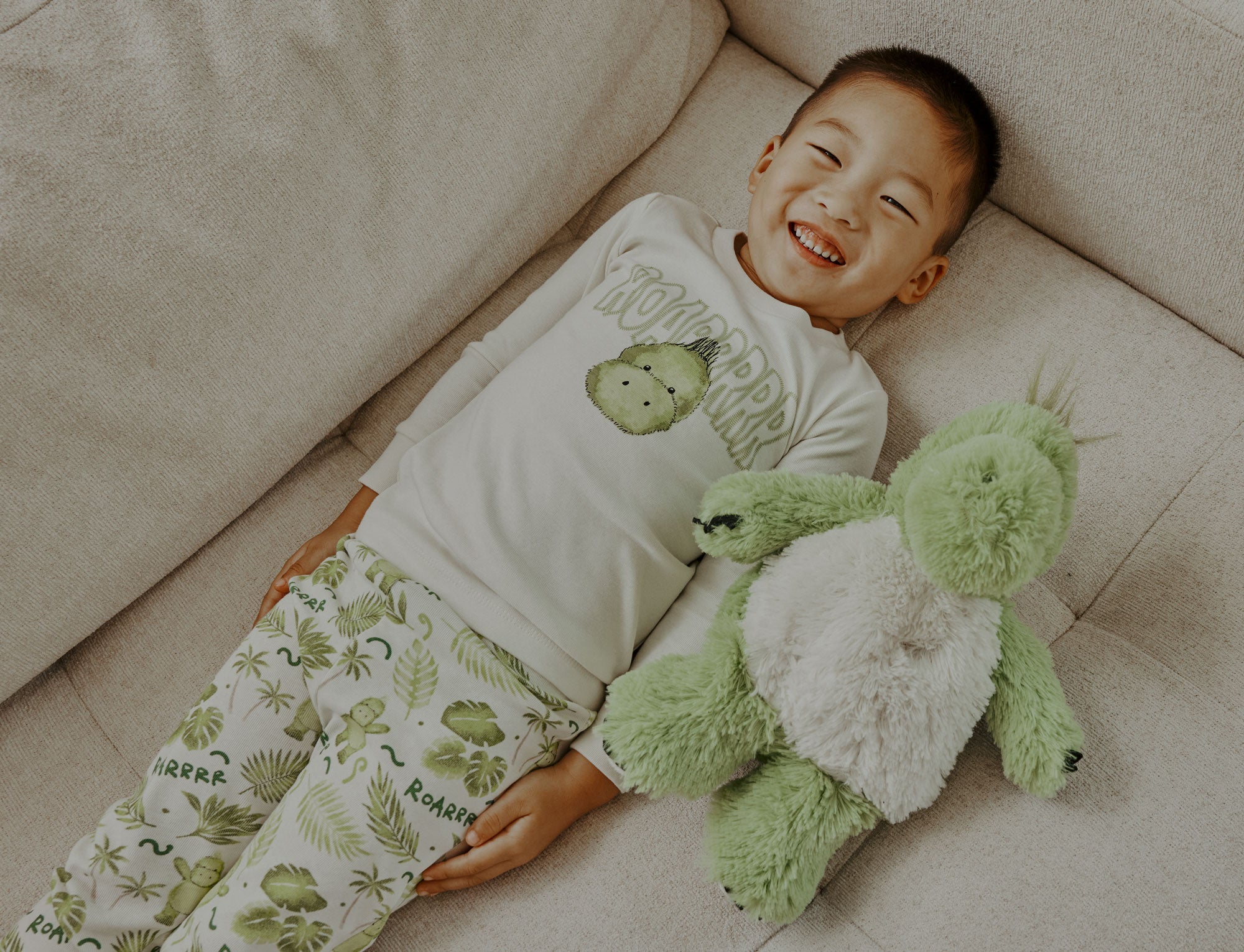 Child smiling laying down next to Weighted, Scented, Cozy Dinosaur Warmies.