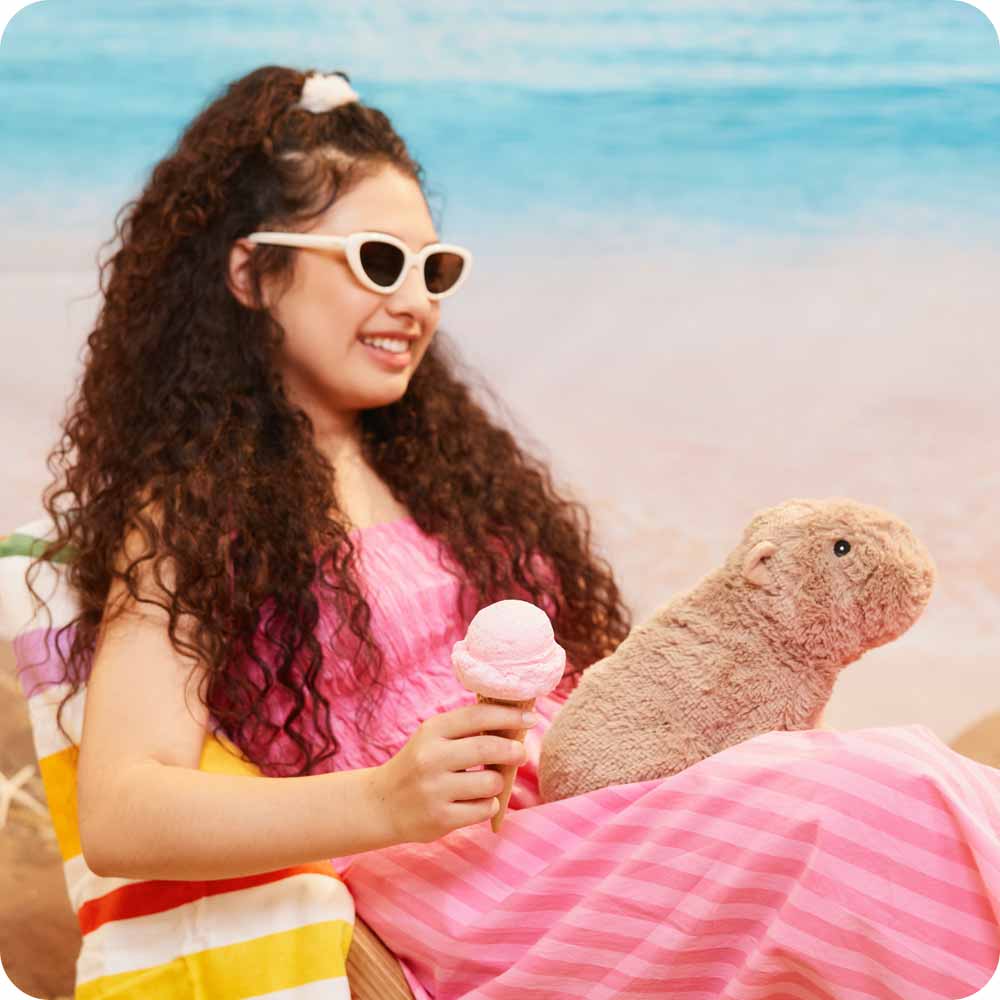 Young teenage girl holding an icecream while sitting on the beach with a Capybara Warmies in her lap