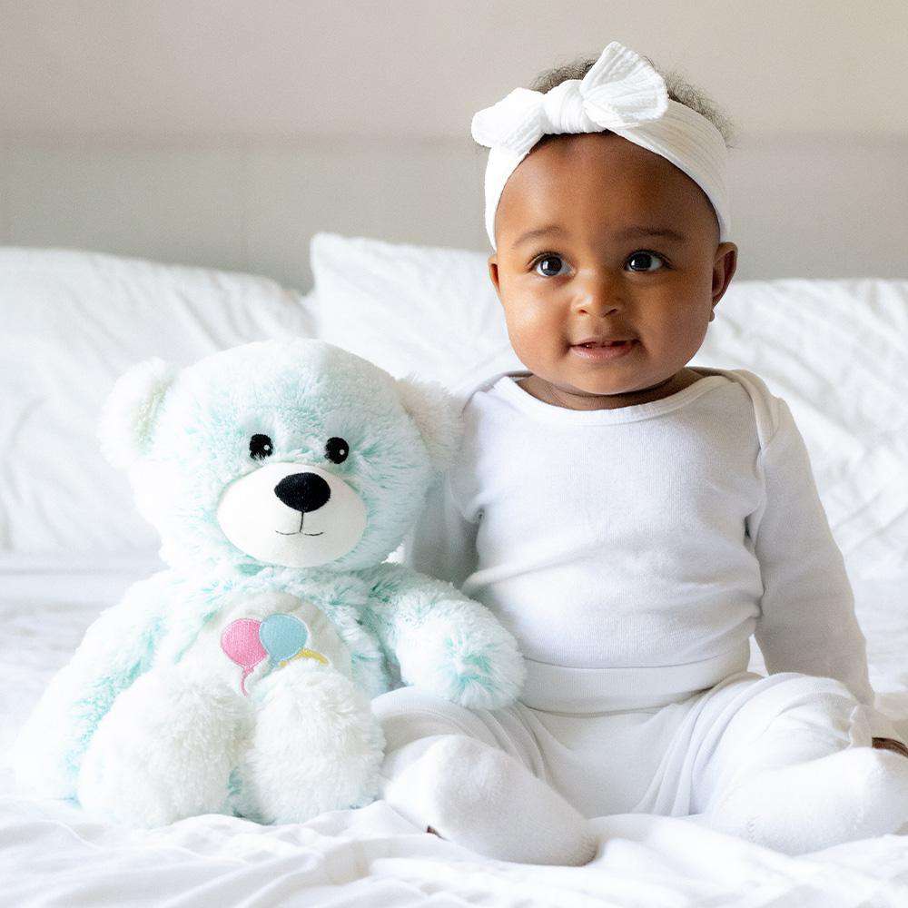 Baby sitting next to Cute Weighted Lavender Scented Celebration Bear Stuffed Animal Heating Pad Warmies