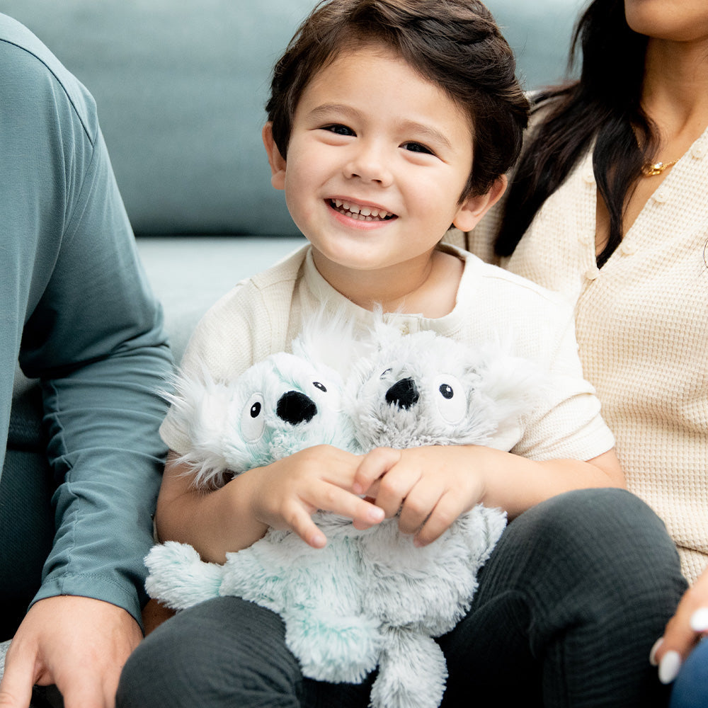 Little boy sitting down and smiling while holding Soft Weighted Lavender Scented Koala Hugs Plush Warmies