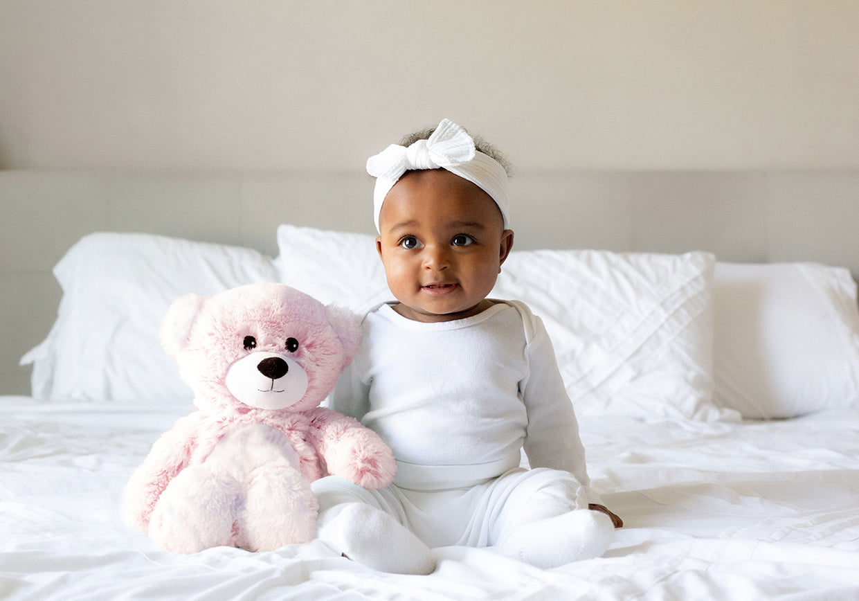 Baby on a white bed with a Pink Marshmallow Bear Warmies.