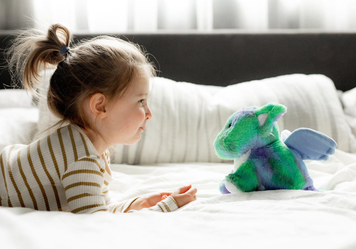 Little girl and blue and green Baby Dragon Warmies laying on a bed.