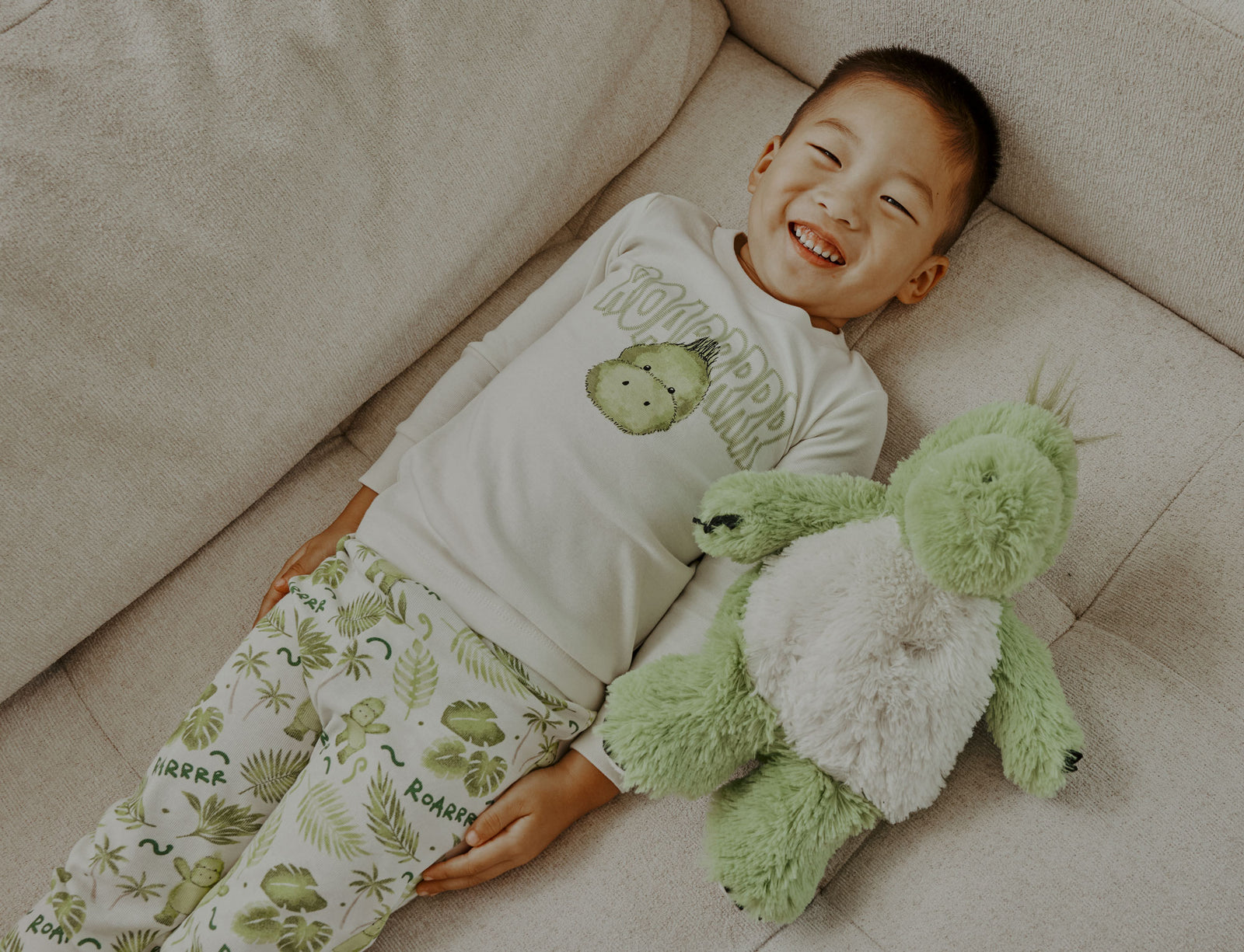 Child smiling laying down next to Weighted, Scented, Cozy Dinosaur Warmies. 