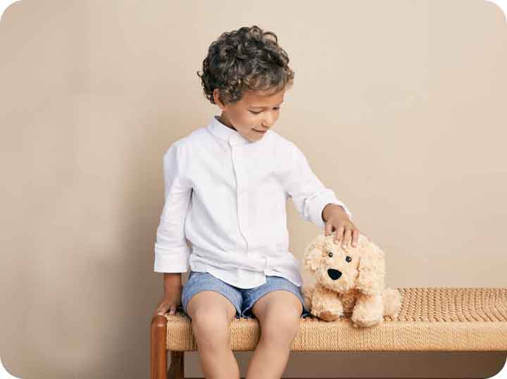 A little boy wearing a white shirt and blue shorts sitting on a brown bench while petting a Warmies Golden Dog by his side. 