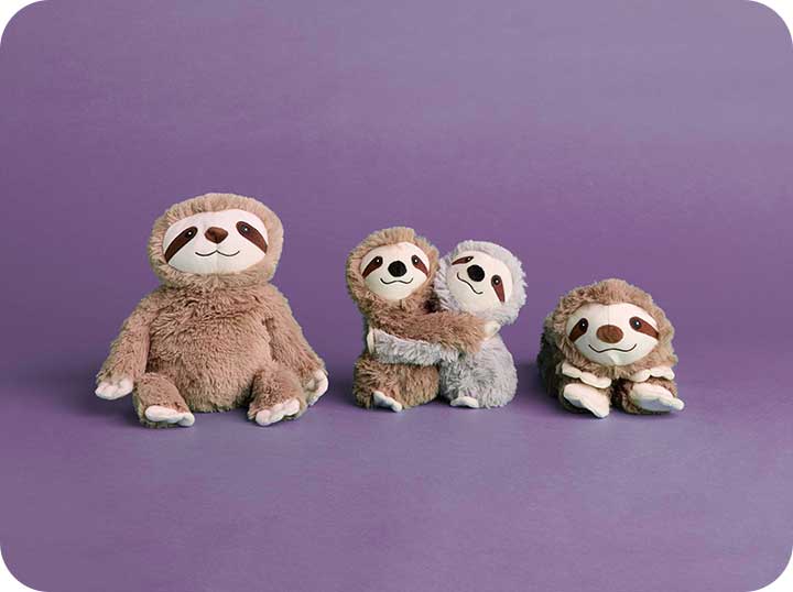 Sloth Warmies, Warmies Hugs and Sloth Animal Neck Wrap sit in a row against a purple background. 