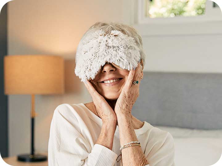 A woman sitting in her bedroom with the Warmies Eye Mask over her eyes, she's smiling and her hands to her cheeks.