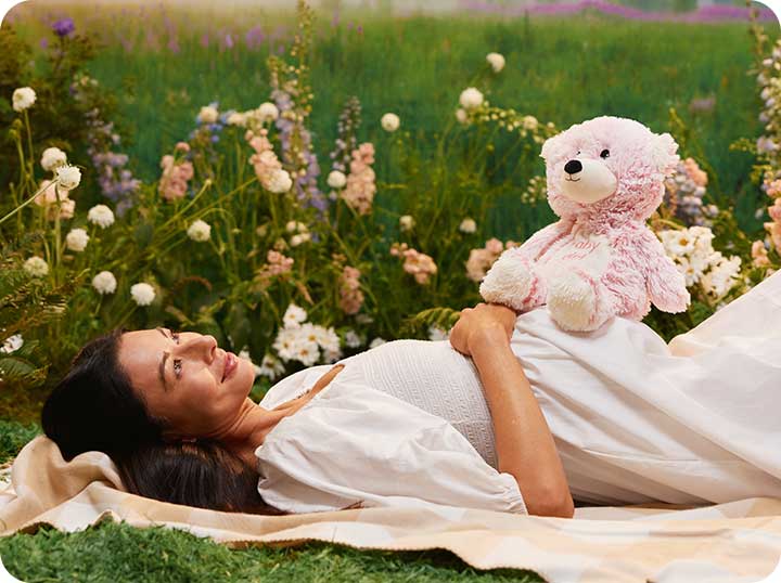 A pregnant woman laying on her back in a garden in a white dress with a Pink Marshmallow Bear Warmies sitting on top of her belly.