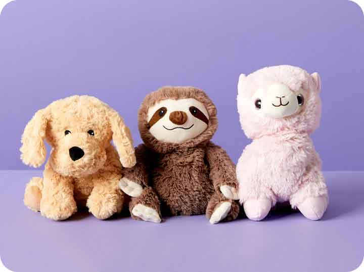 Golden Dog Warmies, Sloth Warmies, Pink Llama Warmies sitting in a row against a purple back ground.