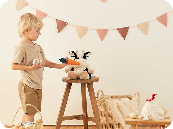 A little boy in a brown outfit feeding a toy carrot to a Warmies Cow, they are surrounded by brown easter baskets with a few eggs and a Chicken Warmies sits on another bench. 