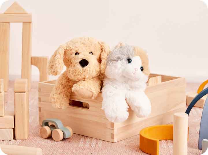 Golden Dog Warmies and Calico Cat Warmies sitting in a wooden toy box surrounded by wooden blocks and toys. 