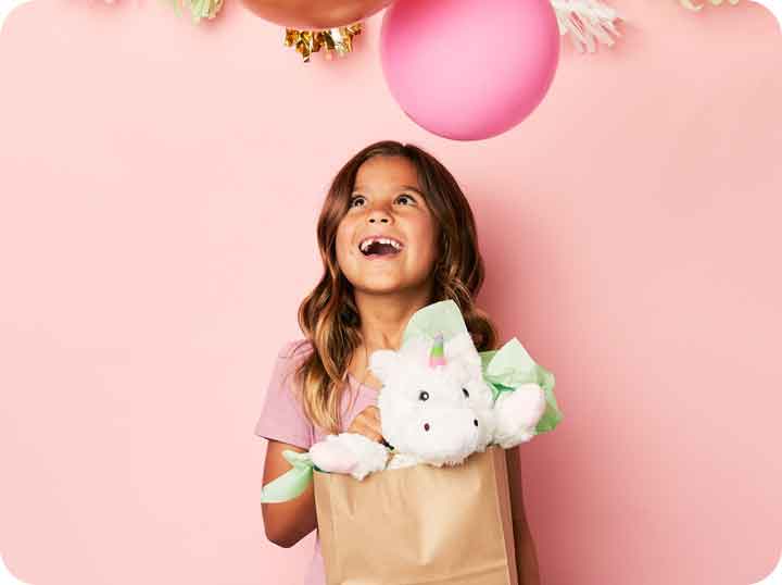 A little girl smiling with balloons above her and she's holding a gift bag with a White Warmies Unicorn popping out from the bag. 