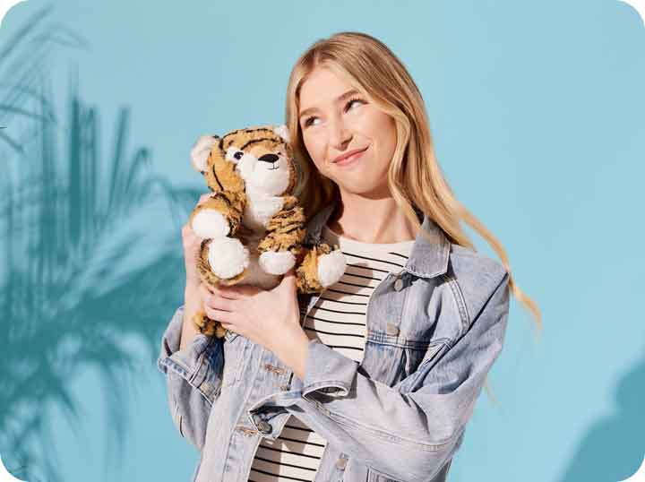 A young girl holding a Tiger Warmies to her cheek with a smirk, against a blue background. 