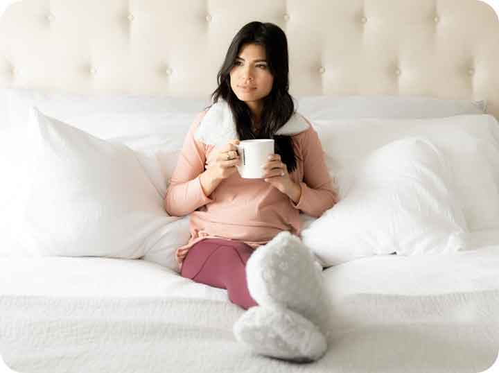 A woman sitting on a white bed holding a cup of coffee and wearing a Warmies neck wrap and Warmies Slippers.